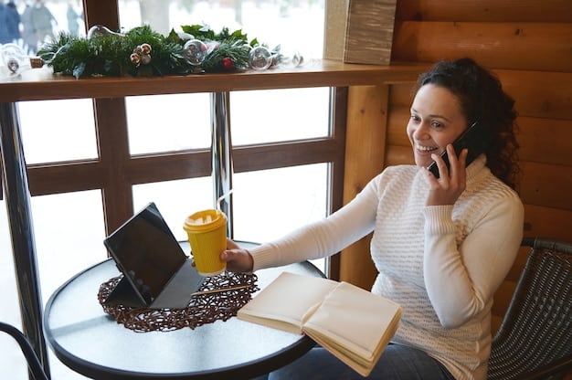 A person working on a laptop in a coffee shop, with their phone displaying a gig work app. Sunlight streams through the window, highlighting the casual and flexible work environment often associated with the gig economy in the US.