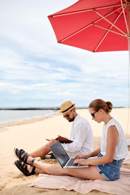 A family of three sitting on a beach in Costa Rica, building a sandcastle together, with a laptop and beach umbrella nearby.