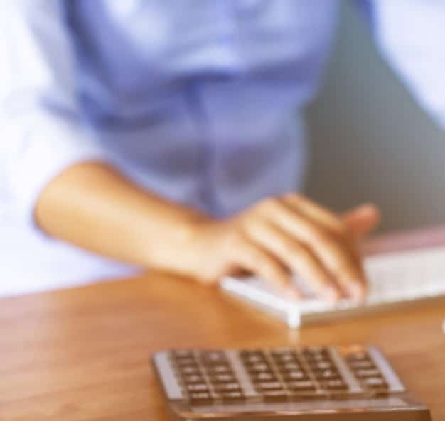 A close-up shot of a freelancer's hands typing on a calculator, with various tax forms and financial documents scattered on the desk around it. The focus is on the act of calculation and financial planning.